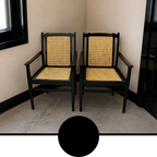 Two matching armchairs with black frames and woven cane seats, placed in a corner room with beige walls and a gray floor. A large black circle overlay appears at the bottom of the frame.