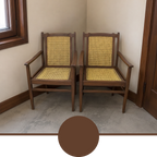 Two wooden dining armchairs with woven cane seats and backs, positioned in a corner of a room with beige walls and a concrete floor. A circular brown color swatch is overlaid at the bottom of the image.